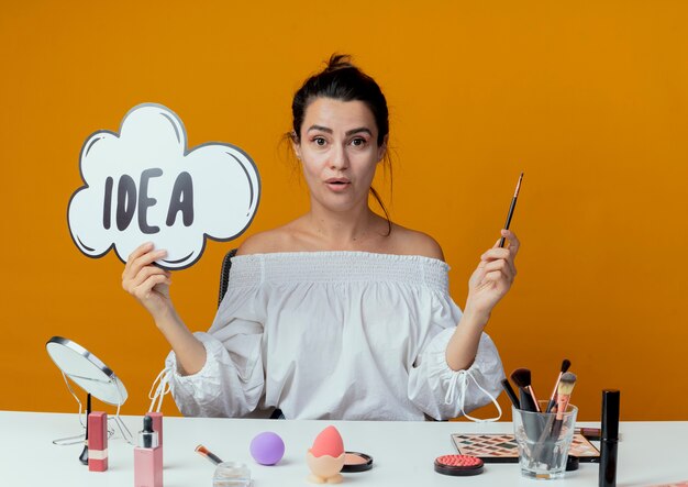 Surpris belle fille est assise à table avec des outils de maquillage tenant une marque d'idée et un pinceau de maquillage isolé sur un mur orange