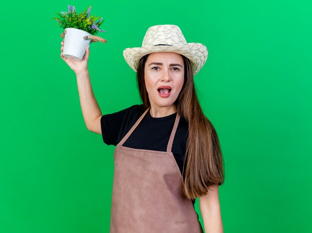 Surpris belle fille de jardinier en uniforme portant chapeau de jardinage soulevant des fleurs en pot de fleurs isolé sur fond vert