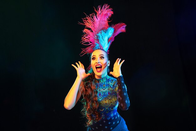 Surpris. Belle jeune femme en carnaval, costume de mascarade élégant avec des plumes sur fond noir en néon.