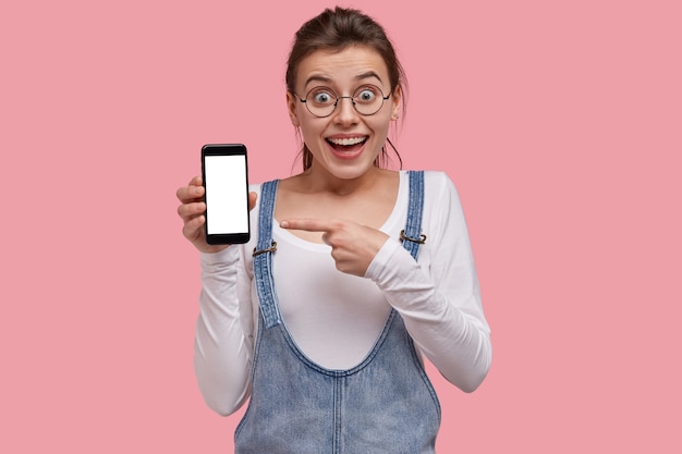 Surpris belle jeune femme indique à l'écran vide du téléphone intelligent, montre un espace pour vos informations, habillé en tenue à la mode