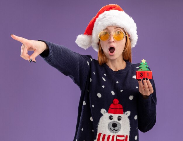 Photo gratuite surpris belle jeune fille portant chandail de noël et chapeau avec des lunettes tenant des points de jouets de noël à côté isolé sur fond violet