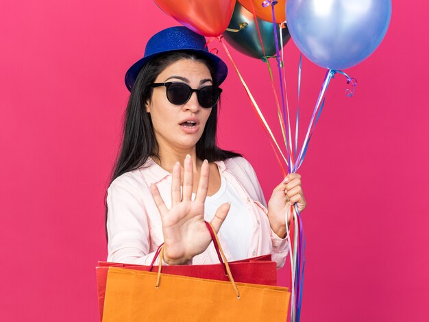 Surpris belle jeune fille portant un chapeau de fête tenant des ballons avec des sacs-cadeaux montrant un geste d'arrêt isolé sur un mur rose