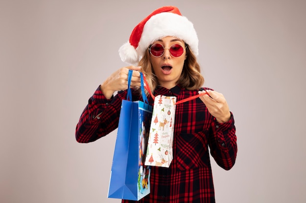 Photo gratuite surpris belle jeune fille portant un chapeau de noël avec des lunettes tenant et regardant des sacs-cadeaux isolés sur fond blanc