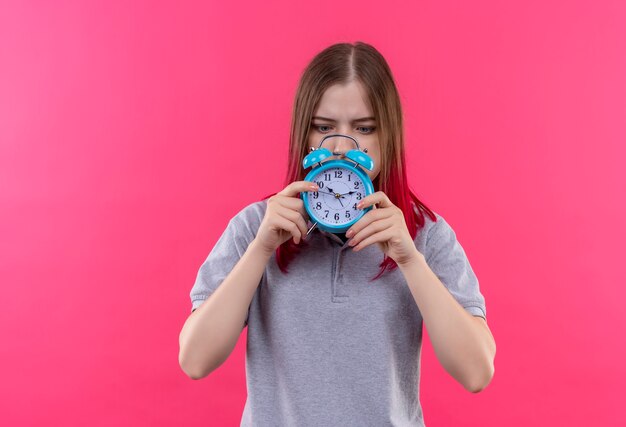 Surpris belle jeune fille portant un t-shirt gris à la recherche d'un réveil dans sa main sur fond rose isolé