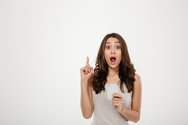 Surpris brunette woman holding smartphone et avoir une idée tout en regardant la caméra sur gris