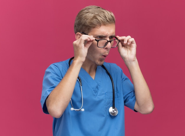 Surpris à côté jeune médecin de sexe masculin portant l'uniforme de médecin avec stéthoscope et lunettes isolé sur mur rose