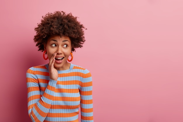 Surpris curieuse femme bouclée garde la main près de la bouche et murmure secret, répand des rumeurs, regarde avec une expression étonnée de côté, habillée avec désinvolture, isolée sur un mur rose, espace vide vide