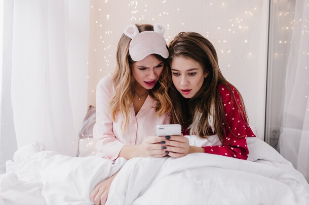 Surpris dames européennes assis dans son lit et regardant l'écran du téléphone. Filles émerveillées en pyjama lisant un message.