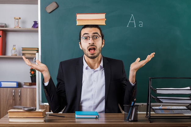 Surpris écartant les mains enseignant portant des lunettes tenant un livre sur la tête assis à table avec des outils scolaires en classe