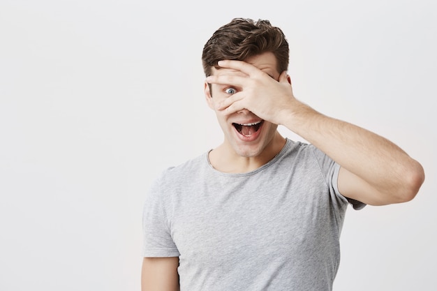 Surpris émotionnel Jeune Homme Européen Ayant Stupéfait Regard Fasciné, Ouvrant La Bouche Largement Et Cachant Le Visage Derrière La Paume, Debout Isolé Sur Fond De Mur De Studio Vide