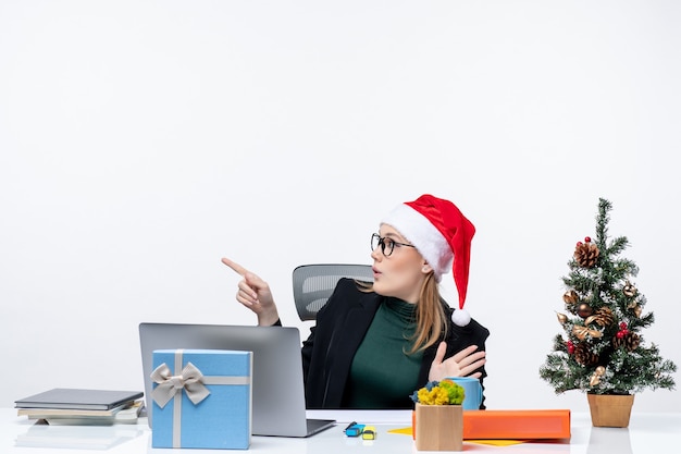 Surpris femme d'affaires pwith son chapeau de père Noël assis à une table avec un arbre de Noël et un cadeau dessus et concentré sur quelque chose avec soin sur fond blanc