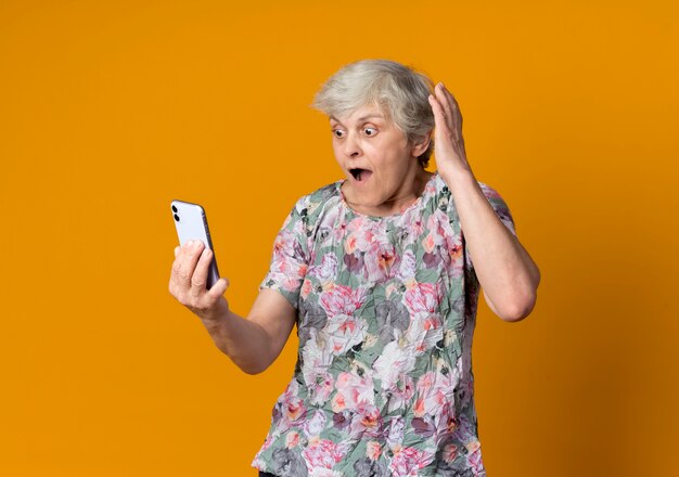 Surpris femme âgée lève la main en regardant téléphone isolé sur mur orange