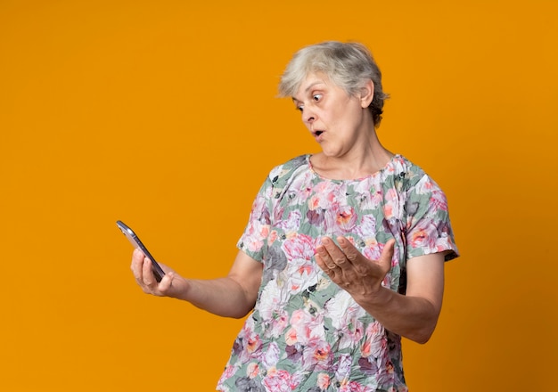 Surpris femme âgée lève la main et regarde le téléphone isolé sur le mur orange