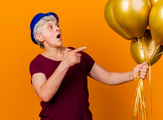 Surpris femme âgée portant chapeau de fête détient des ballons d'hélium à la recherche et pointant sur le côté isolé sur un mur orange avec copie espace