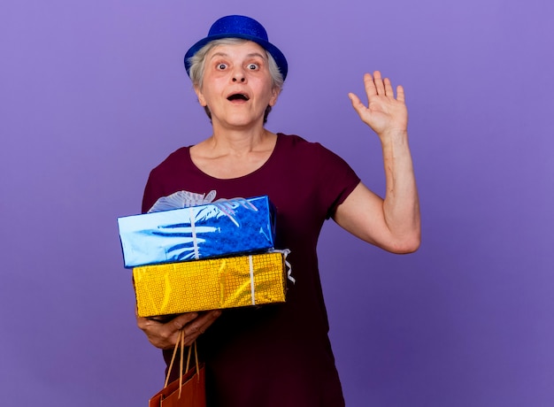 Surpris femme âgée portant chapeau de fête lève la main et détient une boîte-cadeau isolée sur un mur violet avec espace de copie