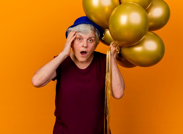 Surpris femme âgée portant chapeau de fête met la main sur le visage et détient des ballons d'hélium isolés sur un mur orange avec copie espace