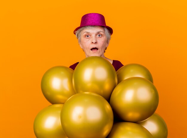 Surpris femme âgée portant chapeau de fête se dresse avec des ballons d'hélium sur orange