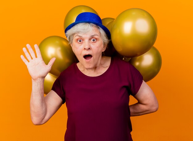 Surpris femme âgée portant chapeau de fête soulève la main et détient des ballons d'hélium derrière isolé sur un mur orange avec copie espace