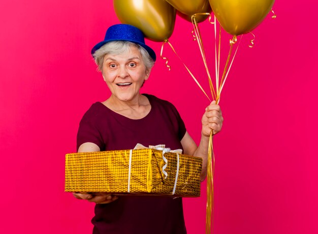 Surpris femme âgée portant chapeau de fête tenant des ballons d'hélium et boîte-cadeau sur rose