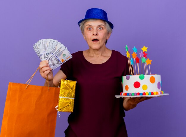 Surpris femme âgée portant chapeau de fête tient un gâteau d'anniversaire boîte-cadeau d'argent