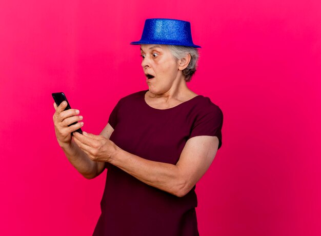 Surpris femme âgée portant chapeau de fête tient et regarde le téléphone sur rose