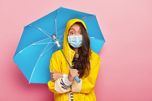 Surpris femme asiatique brune porte un masque médical, étant protégé contre la maladie, porte un imperméable jaune, détient un parapluie