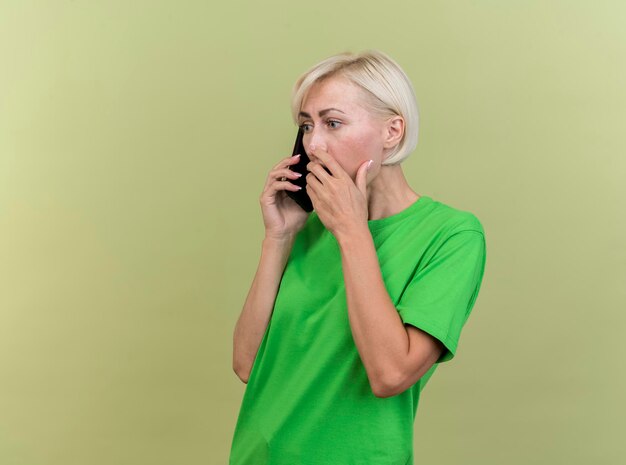 Surpris femme blonde d'âge moyen slave debout en vue de profil parler au téléphone en gardant la main sur la bouche isolé sur mur vert olive avec espace copie