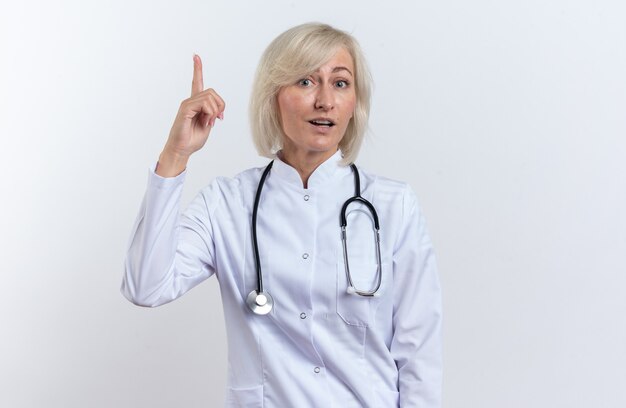 Surpris femme médecin slave adulte en robe médicale avec stéthoscope pointant vers le haut isolé sur fond blanc avec espace de copie