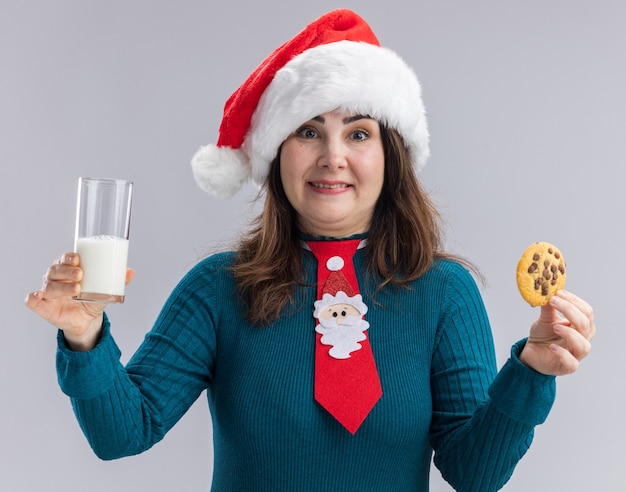 Surpris femme de race blanche adulte avec Bonnet de Noel et cravate de Père Noël tenant un verre de lait et cookie isolé sur fond blanc avec espace copie