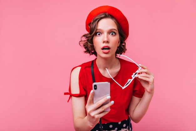 Surpris fille brune posant avec téléphone. Jeune femme bien habillée en béret exprimant son étonnement.