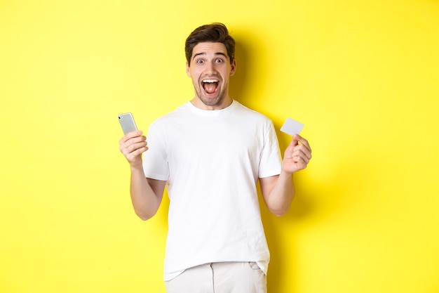 Surpris guy holding smartphone et carte de crédit, achats en ligne le vendredi noir, debout sur fond jaune.