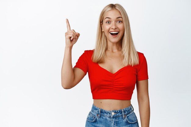 Surpris et heureux femme blonde pointant le doigt vers le haut souriant étonné de vérifier une grande annonce montrant des nouvelles debout sur fond blanc