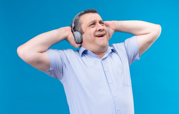 Surpris homme d'âge moyen en chemise rayée bleue portant des écouteurs en profitant de la musique sur fond bleu