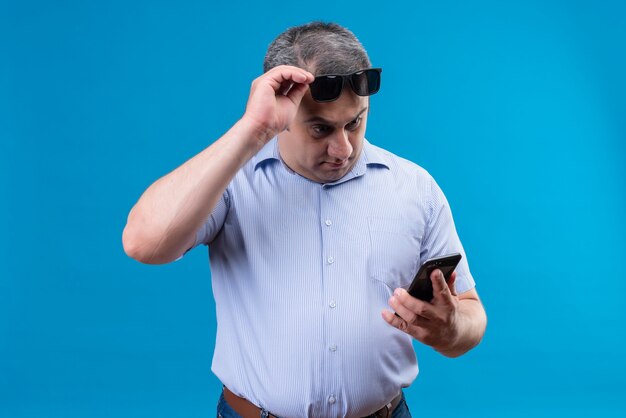 Surpris homme d'âge moyen en chemise rayée bleue portant des lunettes de soleil tenant un téléphone mobile sur fond bleu