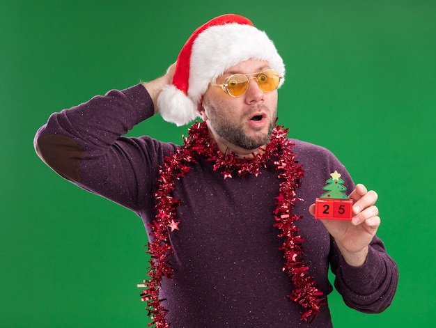 Surpris homme d'âge moyen portant bonnet de Noel et guirlande de guirlandes autour du cou avec des lunettes tenant jouet arbre de Noël