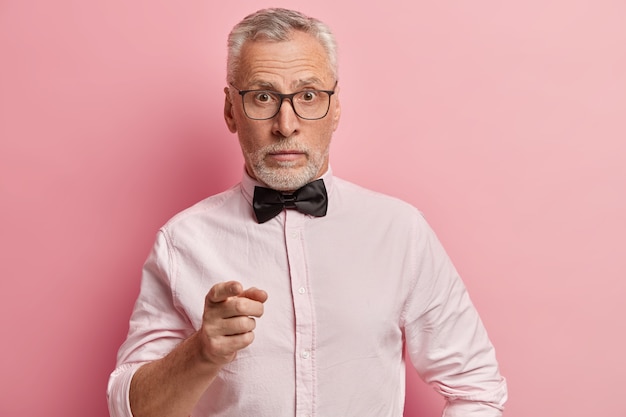 Surpris, un homme âgé porte une chemise élégante avec un noeud papillon noir, des lunettes transparentes, pointe vers la caméra, pose sur fond rose.