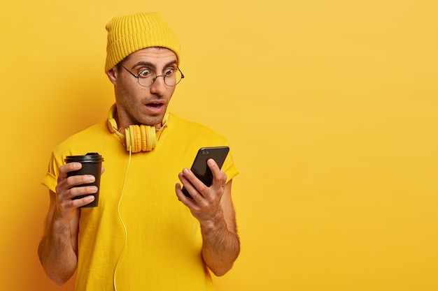 surpris, un homme choqué regarde un smartphone, porte des lunettes transparentes et un chapeau jaune, boit du café à emporter, étonné par de terribles nouvelles sur Internet. Monochrome, couleur jaune