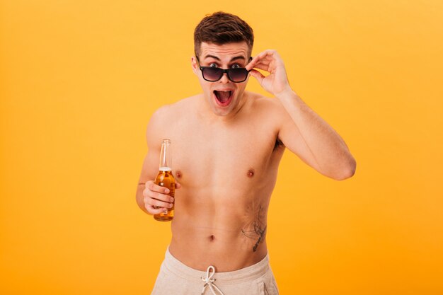 Surpris homme nu en short et lunettes de soleil tenant une bouteille de bière et regardant la caméra par-dessus ses lunettes