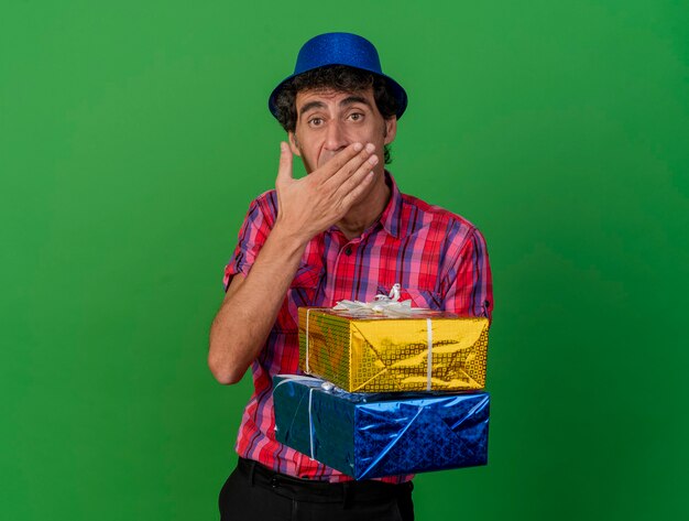 Surpris homme de parti caucasien d'âge moyen portant chapeau de fête tenant des paquets-cadeaux en gardant la main sur la bouche regardant la caméra isolée sur fond vert avec espace copie