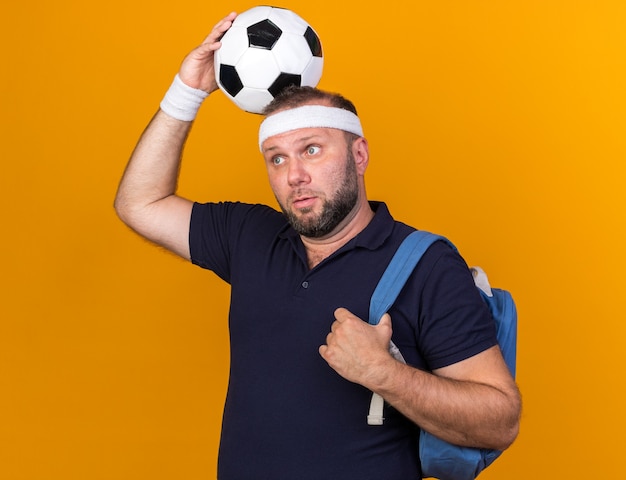 Photo gratuite surpris homme sportif slave adulte portant bandeau sac à dos et bracelets tenant le ballon au-dessus de la tête à côté isolé sur mur orange avec espace de copie