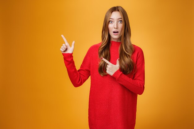 Surpris et impressionné, intrigué, jolie femme rousse mignonne avec des taches de rousseur dans un pull rouge chaud pointant vers le coin supérieur gauche bouche ouverte d'étonnement et regardant la caméra interrogée sur un mur orange