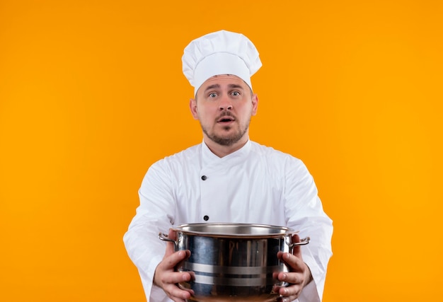 Surpris jeune beau cuisinier en uniforme de chef s'étendant sur la chaudière sur l'espace orange isolé