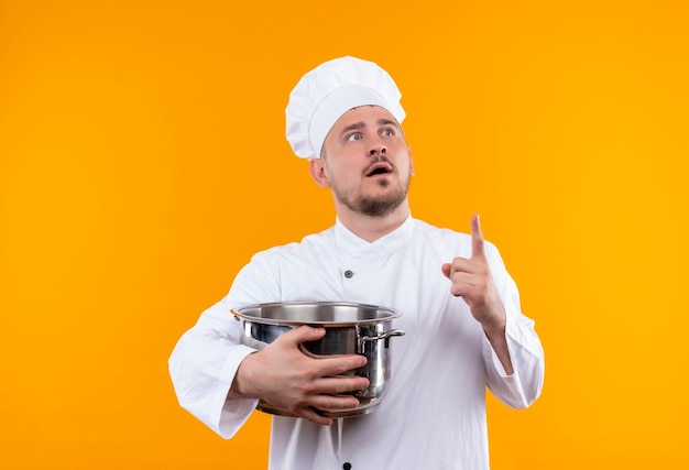 Photo gratuite surpris jeune beau cuisinier en uniforme de chef tenant chaudière et levant le doigt en regardant à droite sur l'espace orange isolé