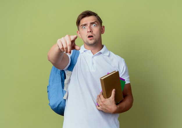 Surpris jeune beau étudiant masculin portant un sac à dos tenant des livres et des points à la caméra isolée sur vert olive