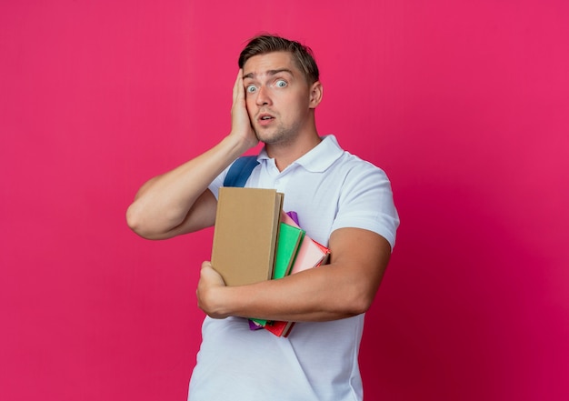 Surpris jeune beau étudiant masculin tenant des livres et mettant la main sur le visage isolé sur rose