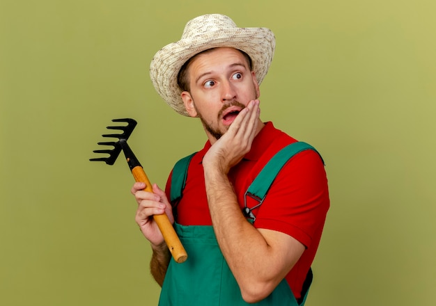 Surpris jeune beau jardinier slave en uniforme et hat holding rake à côté en gardant la main sur le menton isolé sur mur vert olive avec espace copie