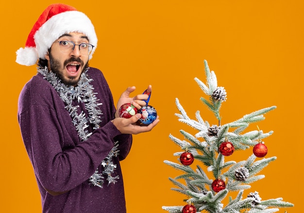 Surpris jeune beau mec debout à proximité de l'arbre de Noël portant un chapeau de Noël avec guirlande sur le cou tenant des boules de Noël isolé sur fond orange