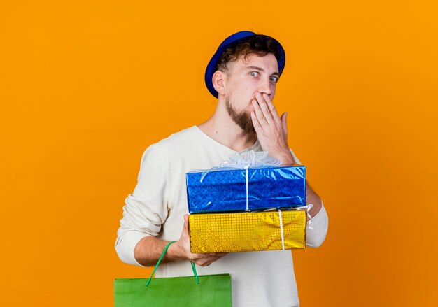 Surpris jeune beau mec de fête slave portant chapeau de fête tenant un sac en papier et des coffrets cadeaux en gardant la main sur la bouche en regardant la caméra isolée sur fond orange avec espace de copie