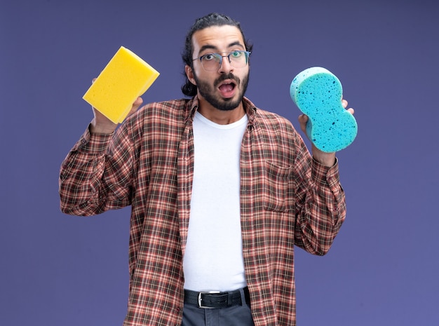 Surpris jeune beau mec de nettoyage portant des t-shirt tenant des éponges isolé sur mur bleu