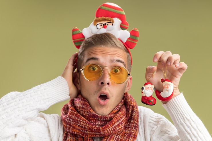 Photo gratuit surpris jeune beau mec portant bandeau et écharpe du père noël tenant des ornements de noël du père noël regardant la caméra touchant la tête isolée sur fond vert olive